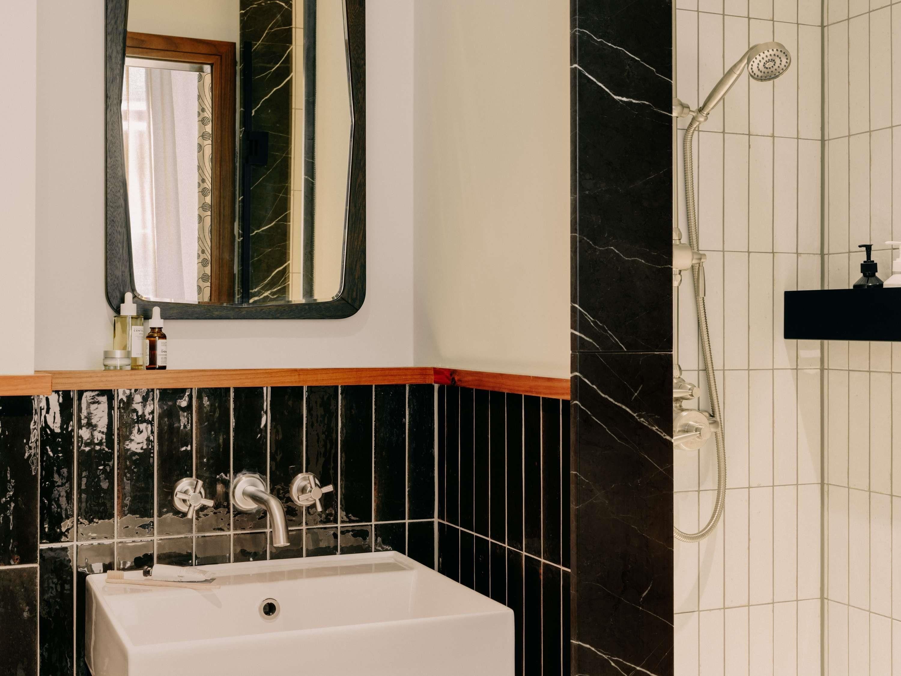 Um banheiro com pia branca, espelho, chuveiro e parede revestida de azulejos pretos.