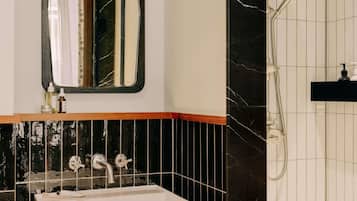 Um banheiro com pia branca, espelho, chuveiro e parede revestida de azulejos pretos.