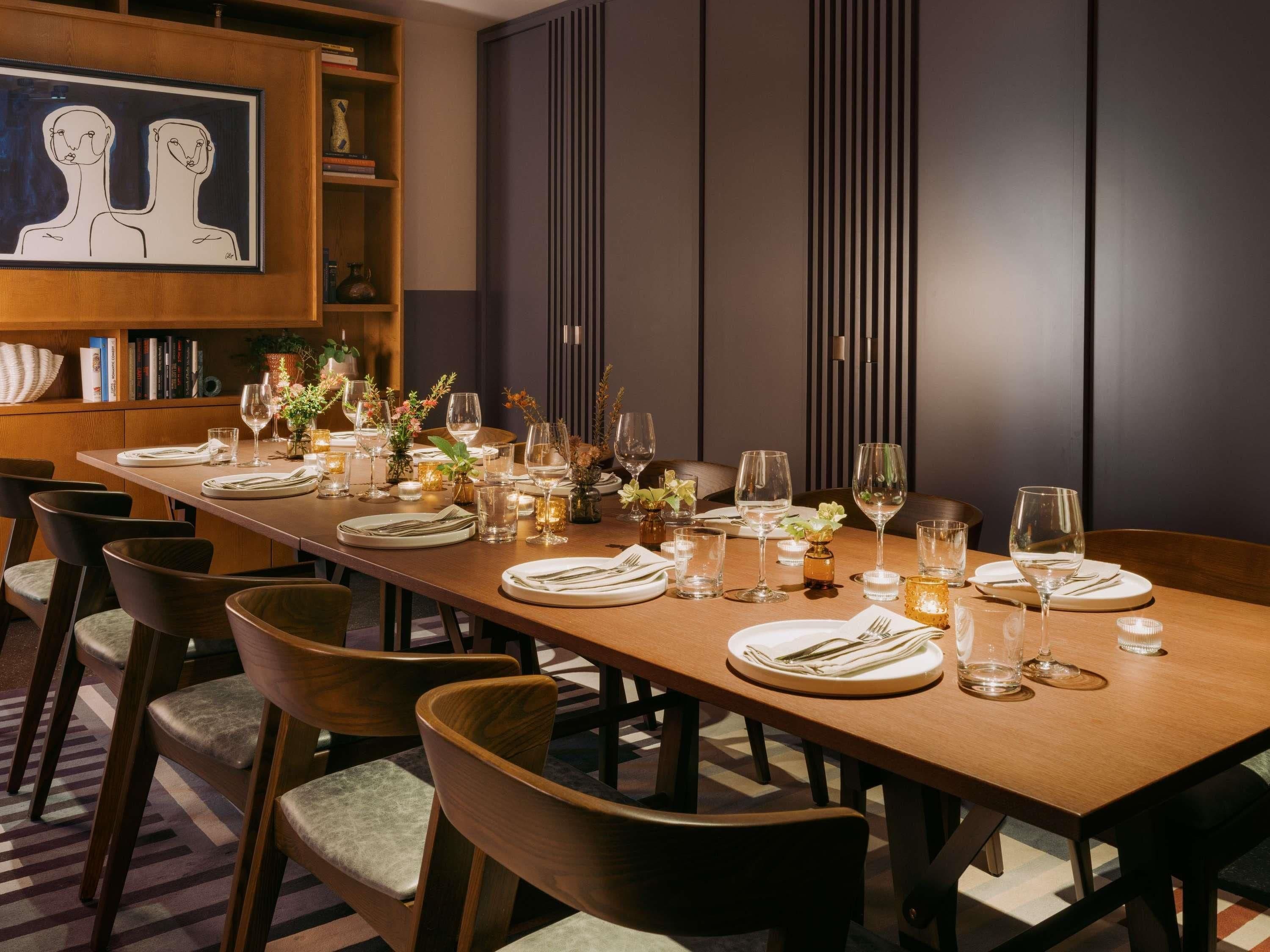 A long dining table set with chairs, candles, and glassware, in a room with a bookshelf and a framed artwork on the wall.