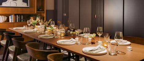 A long dining table set with chairs, candles, and glassware, in a room with a bookshelf and a framed artwork on the wall.