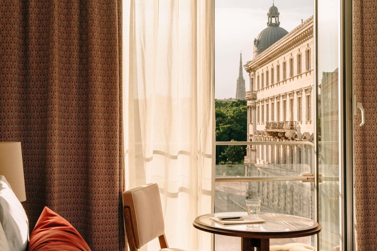 Une chambre d’hôtel avec un balcon, une vue sur un bâtiment historique et une table avec un verre d’eau.