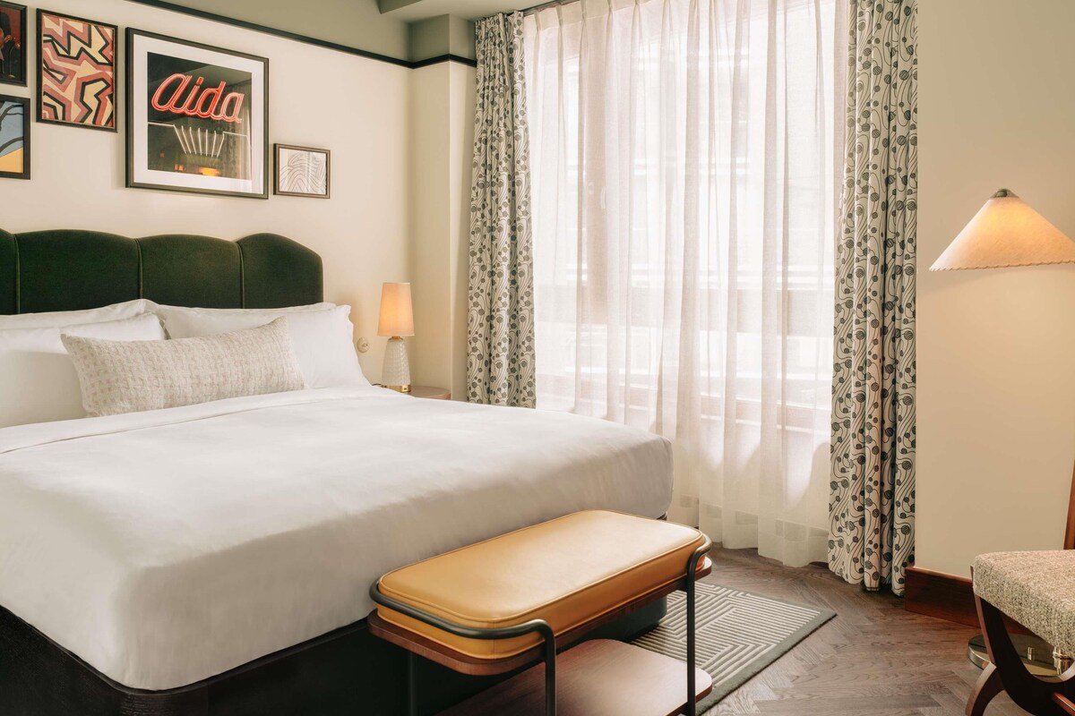 A neatly made bed with a headboard, a bedside table with a lamp, a bench, and a chair.