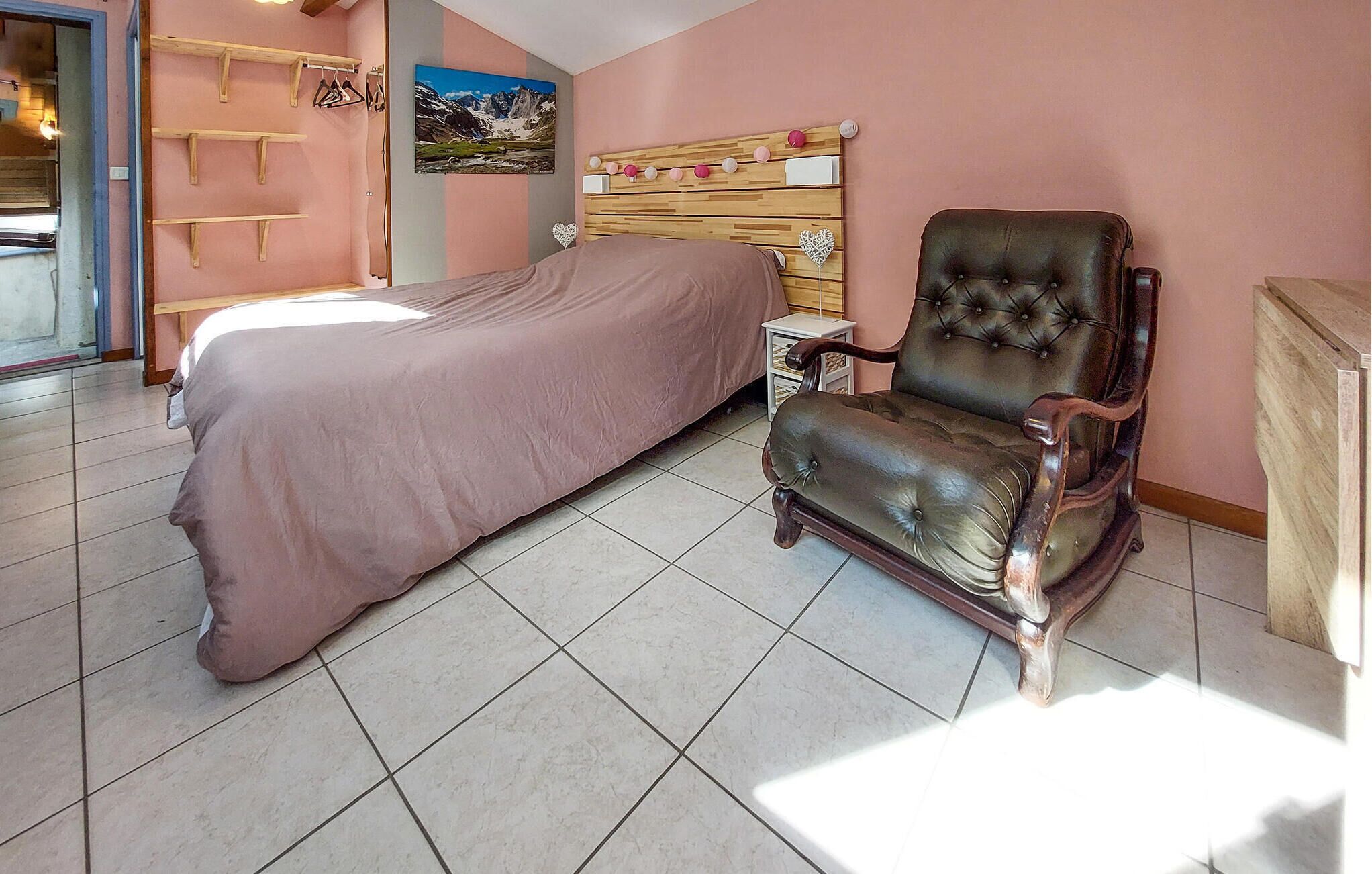 A bedroom with a bed, a leather armchair, a wooden headboard, a small bedside table, and a wall-mounted coat rack.
