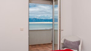 A room with a bed, a chair, and a sliding glass door leading to a balcony with a view of mountains and a beach. - Apartments Palaic (Vir)
