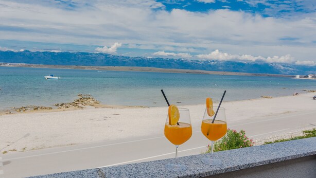 Two glasses of orange juice with straws on a balcony overlooking a beach with a boat in the water and mountains in the background. - Apartments Palaic (Vir)