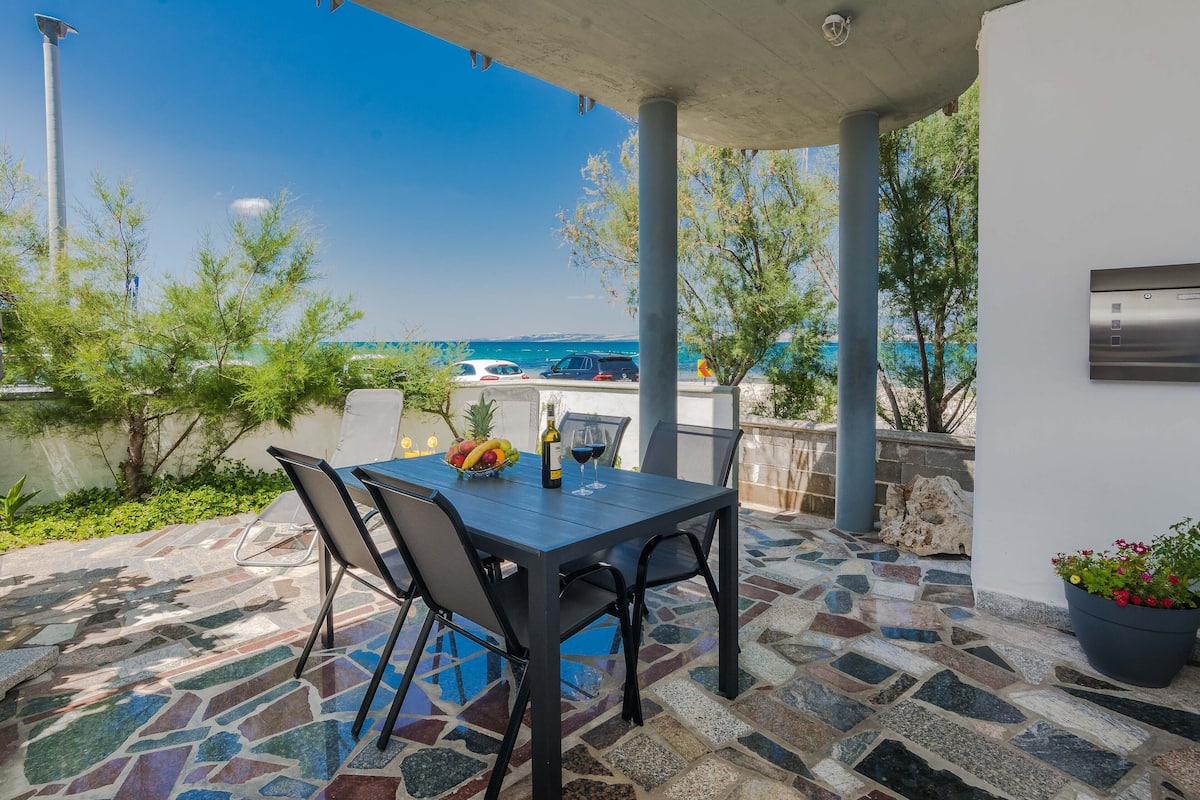 An outdoor patio with a stone-tiled floor, a table with chairs, a bottle of wine, and a bowl of fruit.