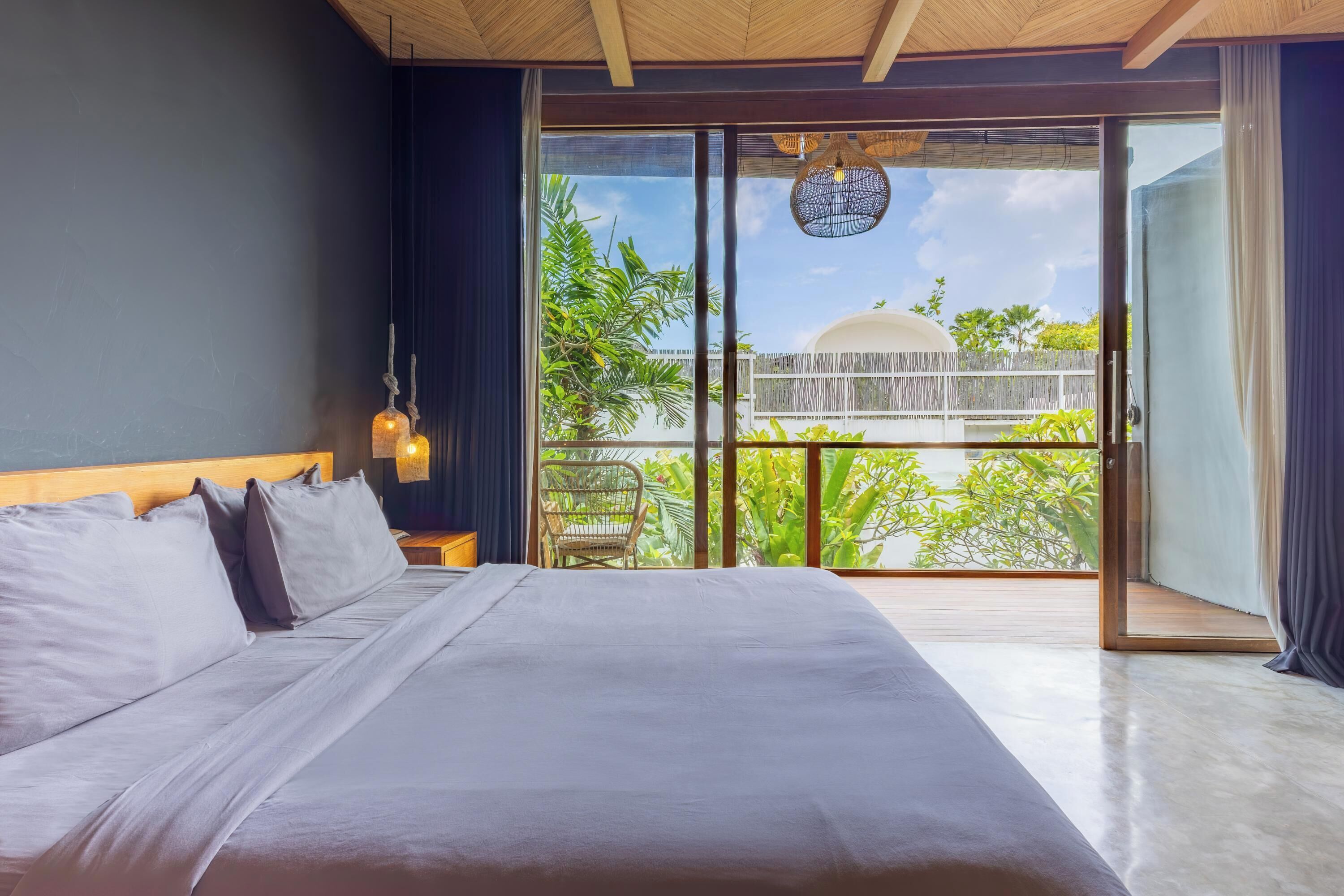 Une chambre avec un grand lit, une table de chevet, un luminaire suspendu et une vue sur un balcon agrémenté de plantes et d’une clôture.