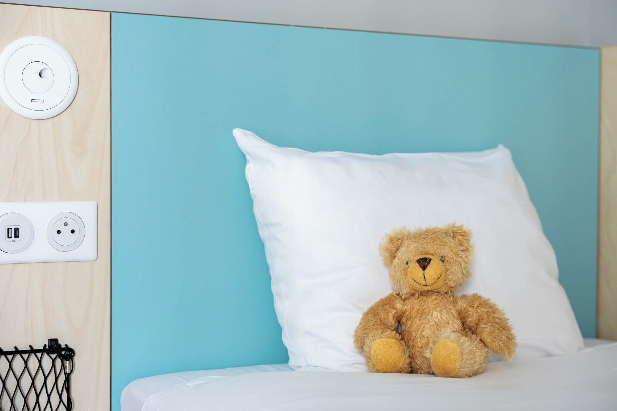 Ein Bett mit einem weißen Kopfkissen und einem Teddybär. An der Wand befinden sich eine Steckdose und ein USB-Anschluss.