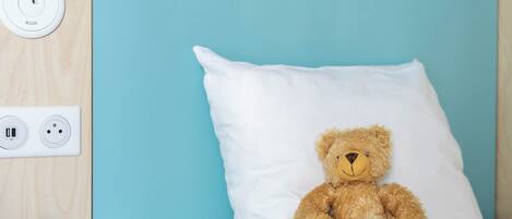 A bed with a white pillow and a teddy bear. There is a power outlet and a USB port on the wall.