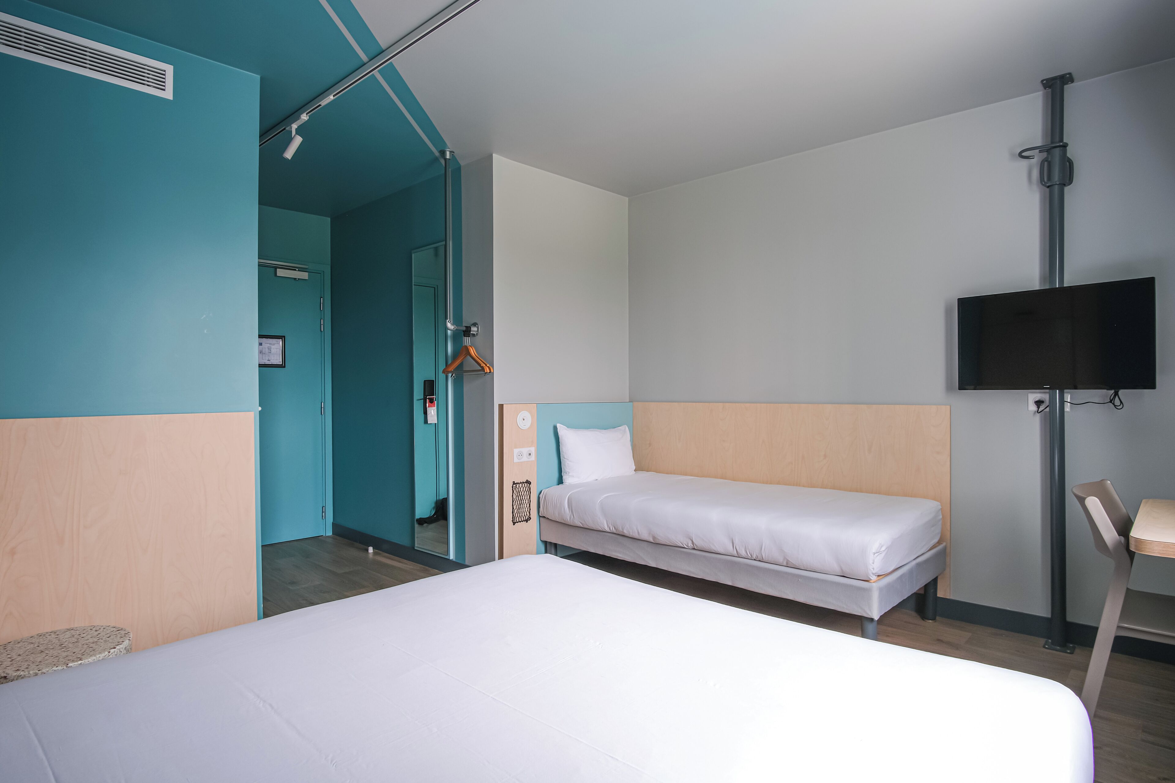 A hotel room with a large bed, a smaller bed, a TV mounted on the wall, and a wooden chair.