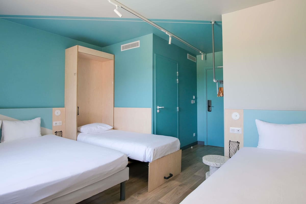 Habitación de hotel con dos camas individuales, un armario de madera, baño con ducha y una puerta que da a otra habitación.