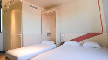 A hotel room with two single beds, a wardrobe, a small table, and a wall-mounted light fixture.
