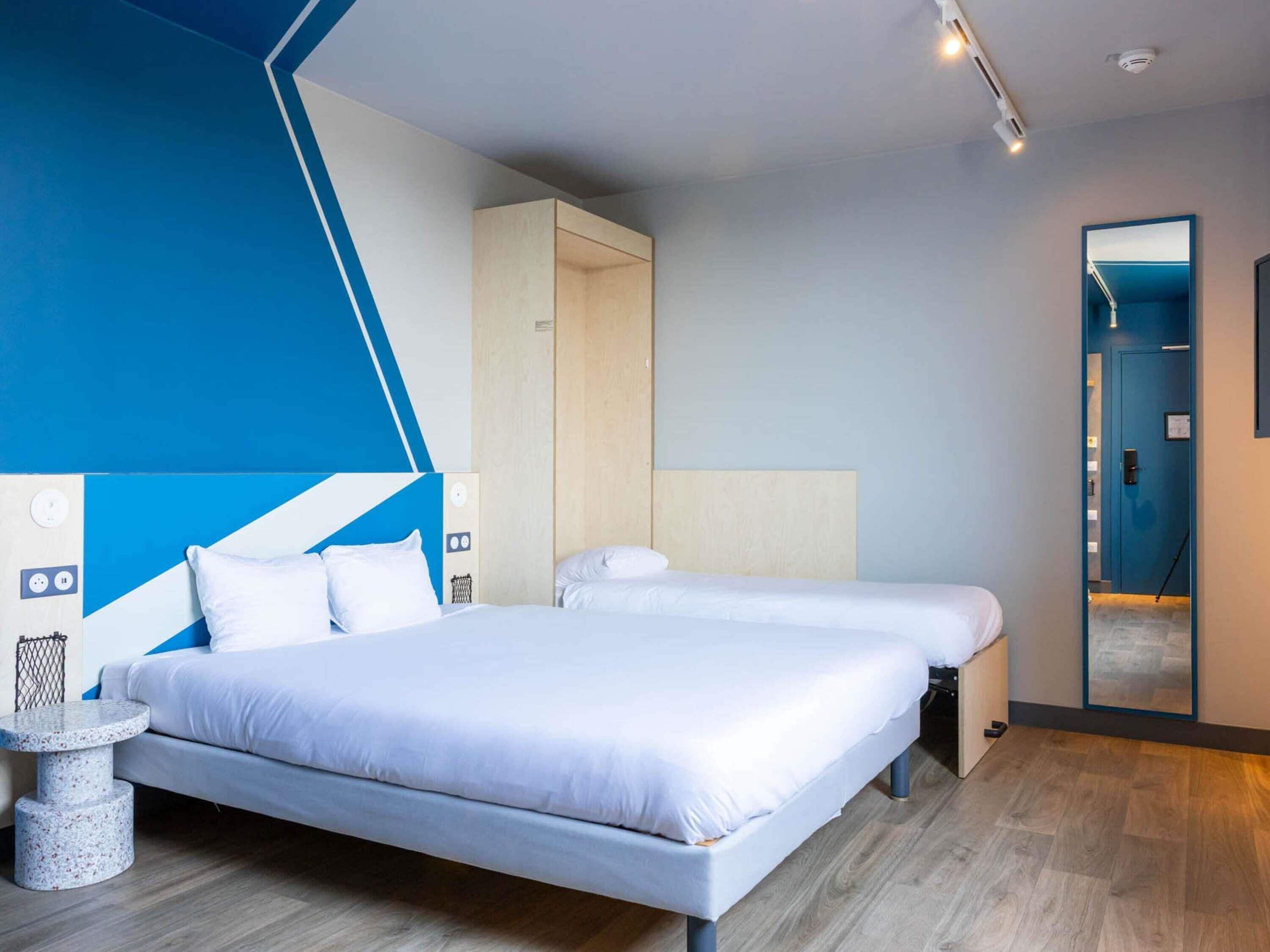 A modern hotel room with two beds, a blue accent wall, a wooden floor, and a small round table.
