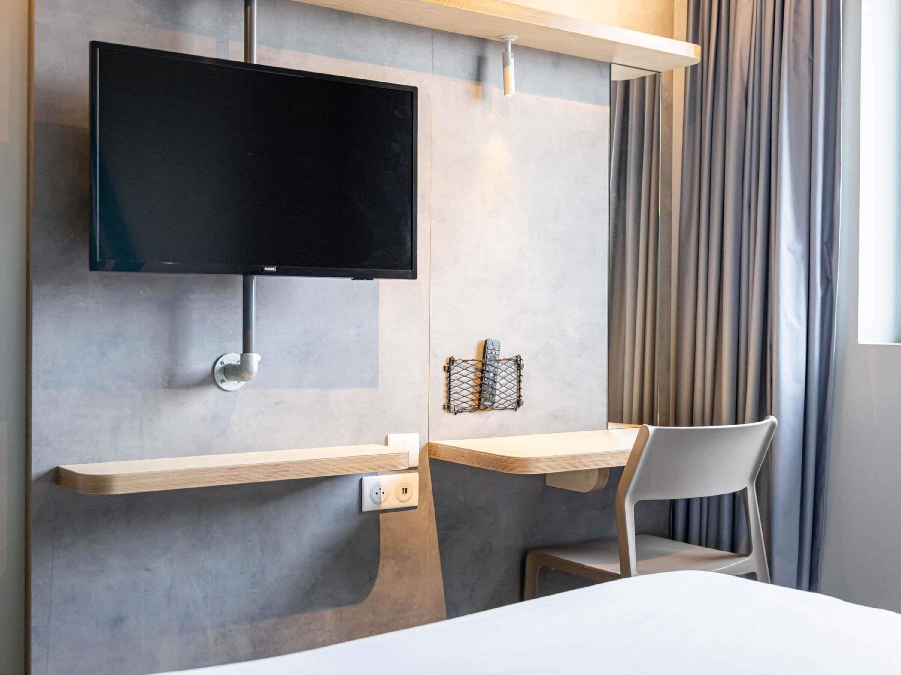 Et moderne hotelværelse med et fladskærms-tv på væggen, et træskrivebord med stol og en stikkontakt.