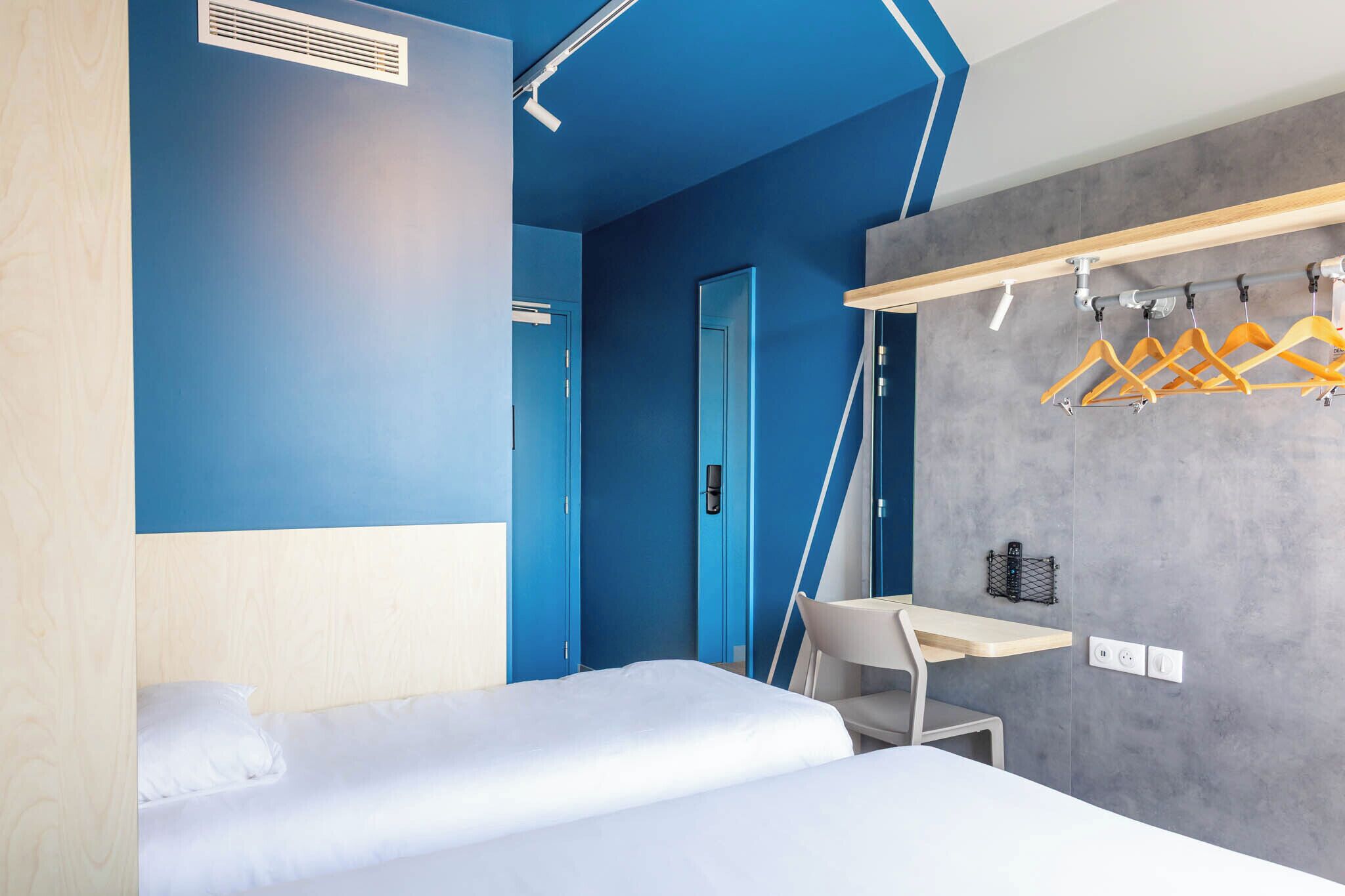 Una habitación de hotel moderna con una cama, un escritorio y una silla. Hay un perchero con ganchos y una pequeña mesa con una silla.