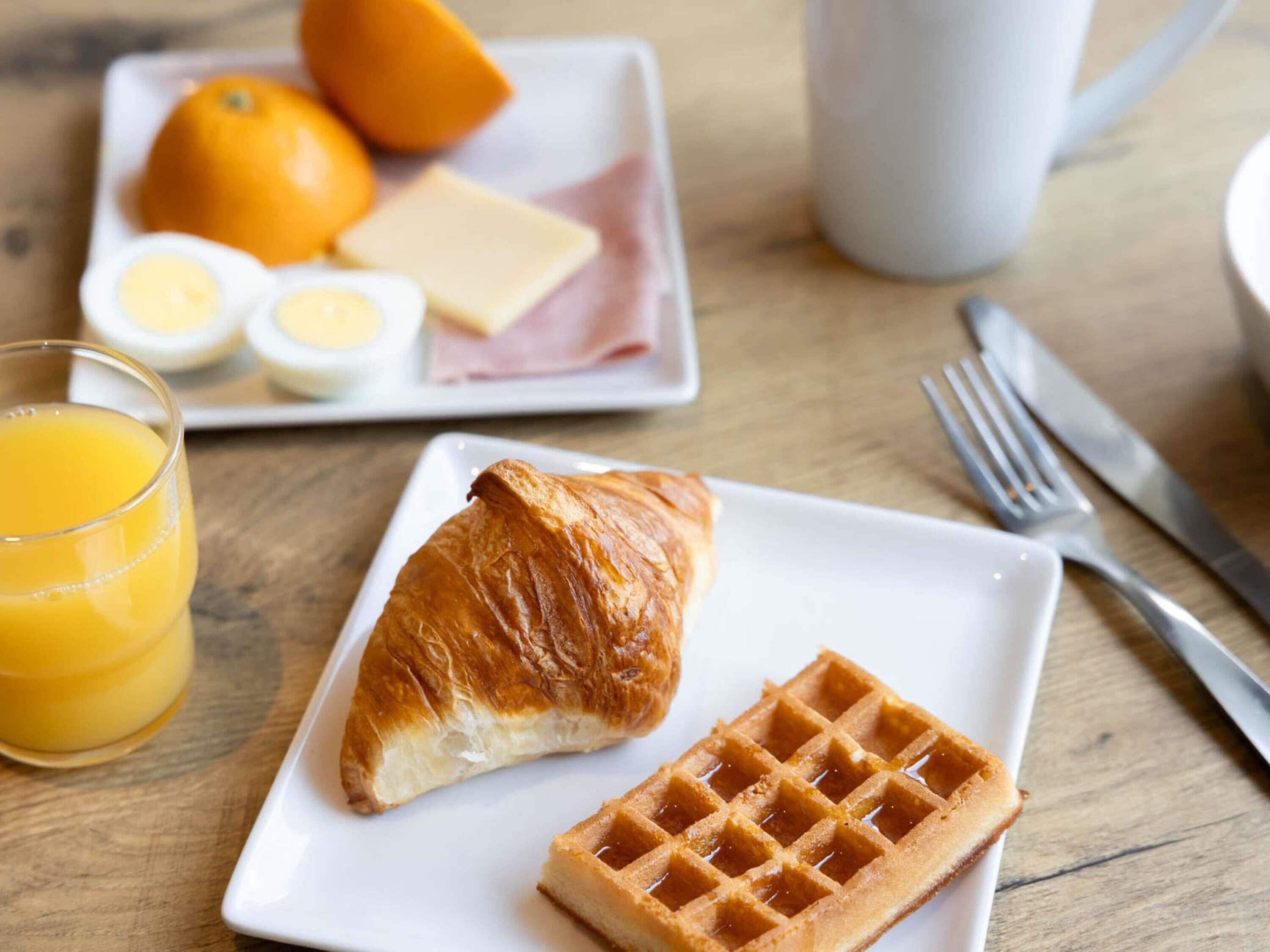 Prato de café da manhã com croissant, waffle, laranja, ovos, queijo e presunto.