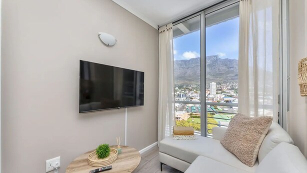 A modern living room with a flat-screen TV mounted on the wall, a round wooden table with a remote and a small plant, a white sofa with a cushion, and a large window offering a city view. - Picturesque 2BD Apartment With Table Mountain View (Cape Town)