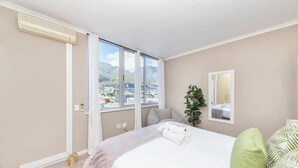 A bedroom with a large bed, a view of a cityscape through a window, a plant, and a mirror on the wall. - Picturesque 2BD Apartment With Table Mountain View (Cape Town)