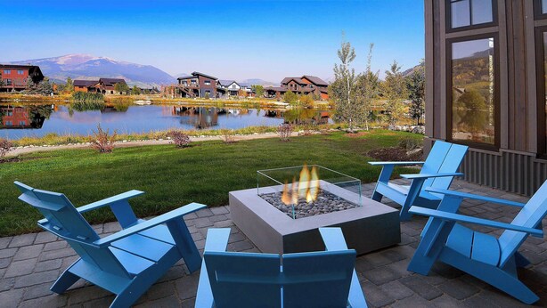 A patio with Adirondack chairs and a fire pit, overlooking a lake and buildings.
