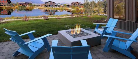 Une terrasse avec des fauteuils Adirondack et un foyer, offrant une vue sur un lac et des bâtiments.