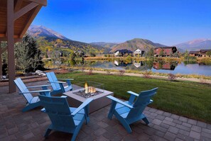 Un patio junto al lago con mobiliario exterior y una fogata, rodeado de montañas y edificios residenciales.