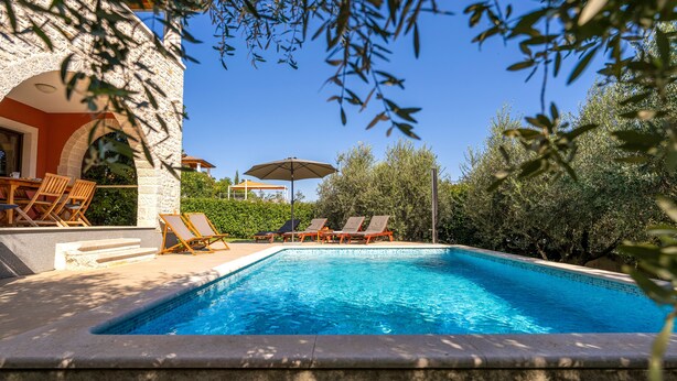 Una piscina con sillas de descanso, una mesa con sillas y un edificio con fachada de piedra.