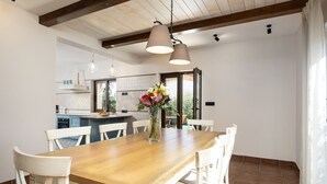 Un comedor con una mesa de madera y sillas blancas, un florero con flores e iluminación colgante.