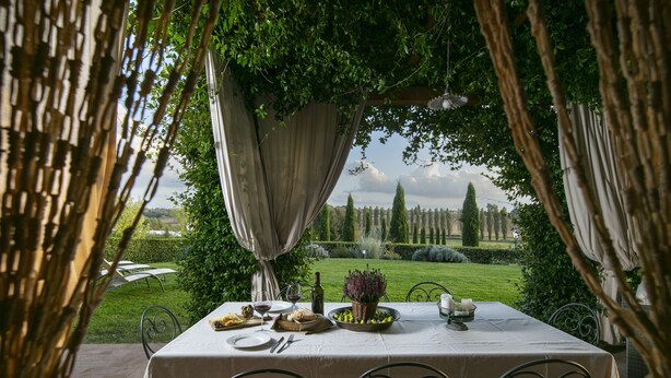 An outdoor dining area with a table set for two, surrounded by metal chairs, a white tablecloth, and a view of a manicured garden and distant trees. - Exclusive luxury property with private pool in Siena countryside Borgo la Chiusa (Siena)