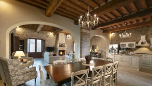 A spacious living area with a wooden dining table, white chairs, and a chandelier. There is a fireplace, stone walls, and a kitchen area with white cabinets. - Exclusive luxury property with private pool in Siena countryside Borgo la Chiusa (Siena)