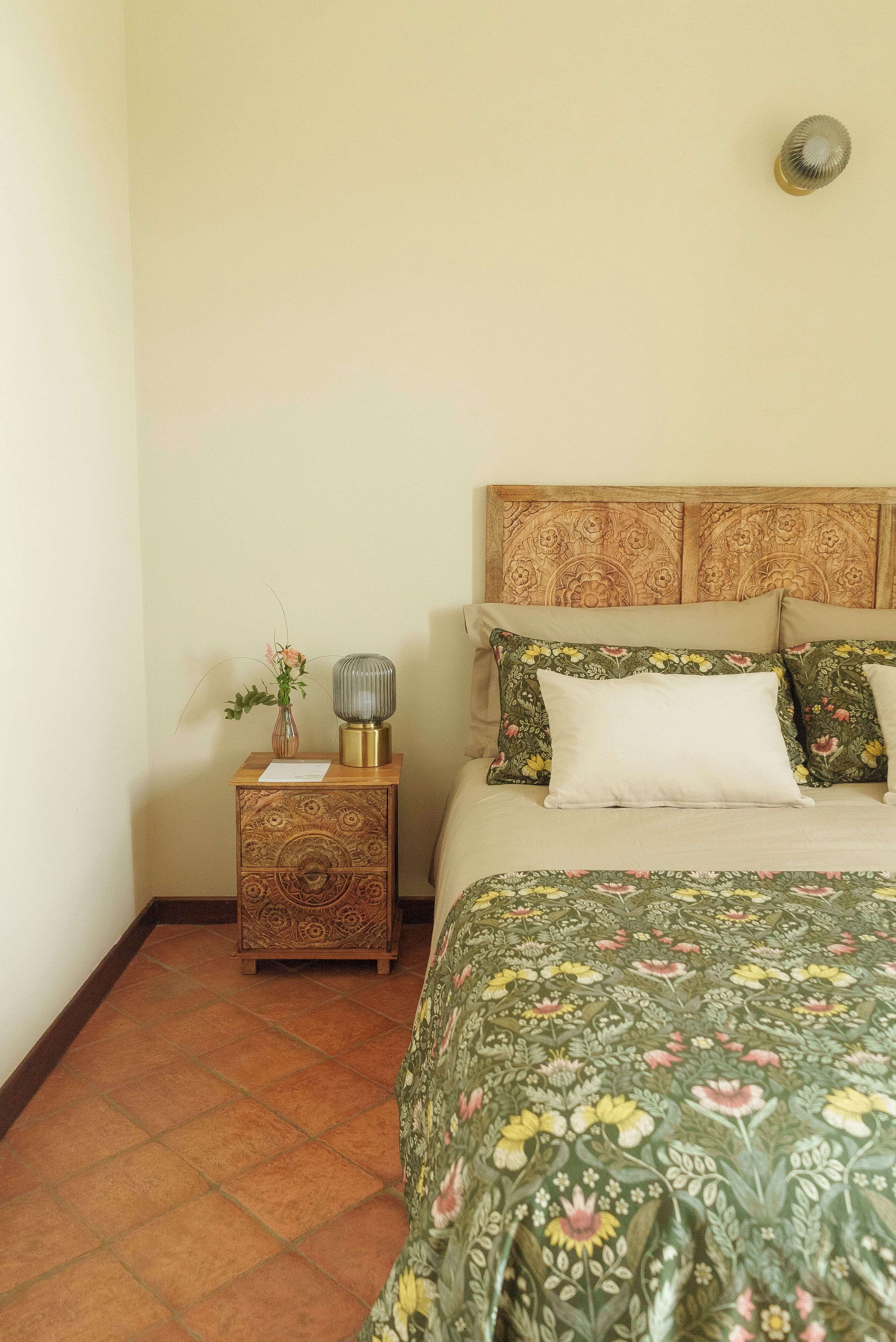 A bedroom with a floral-patterned bedspread, a wooden nightstand with a vase of flowers, and a decorative wall-mounted light.