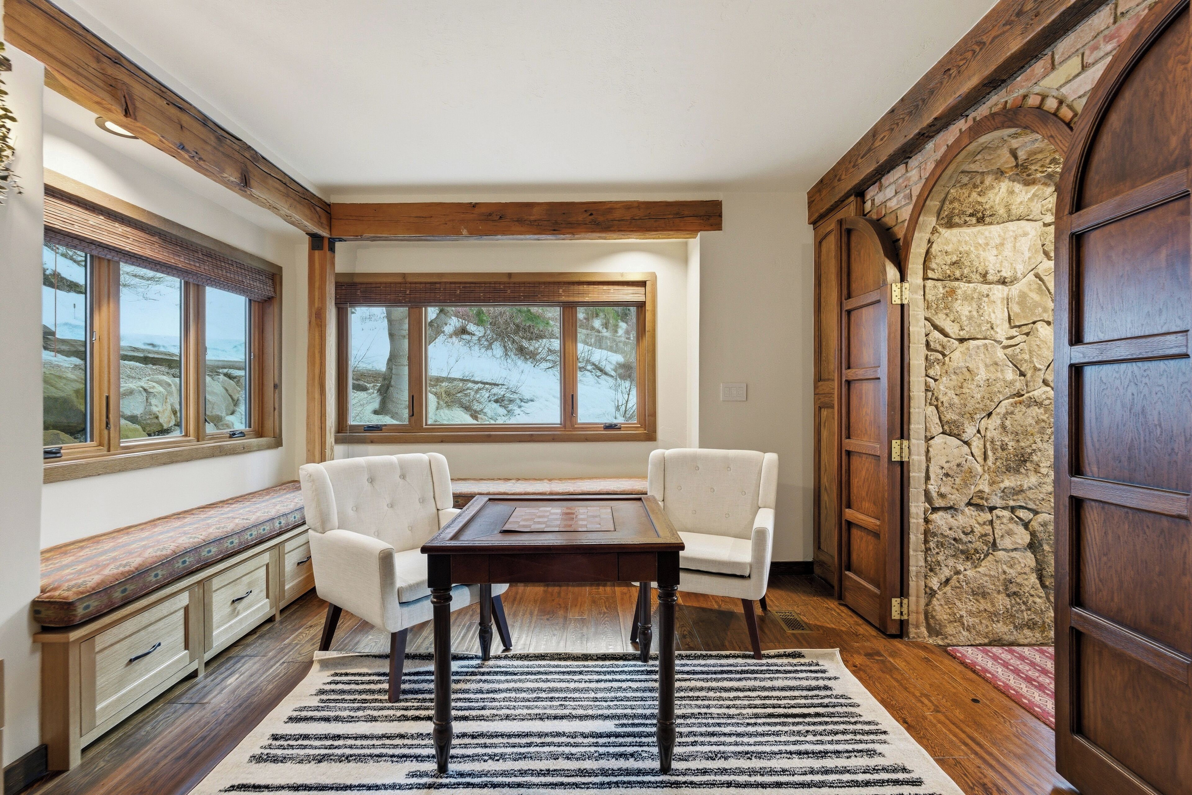 A dining area with a table and chairs, a bench with storage, large windows, and a stone wall.
