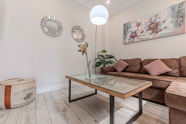 A modern living room with a glass coffee table, a brown leather sofa, a large mirror, and a floral painting on the wall.
