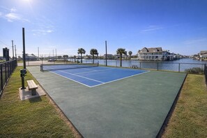 A tennis court with a water view, surrounded by a fence and benches.