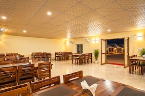 A spacious dining area with multiple wooden tables and chairs, a ceiling with a geometric pattern, and a view of the outside at night. -  Barkha Desert Safari Camp Sam Sand Dunes (Jaisalmer)