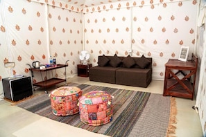 A tented room with a sofa, two colorful ottomans, a fan, a small table with bottles, and a speaker. -  Barkha Desert Safari Camp Sam Sand Dunes (Jaisalmer)