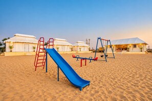 A sandy beach with a blue slide, red and blue swings, and white tents in the background. -  Barkha Desert Safari Camp Sam Sand Dunes (Jaisalmer)