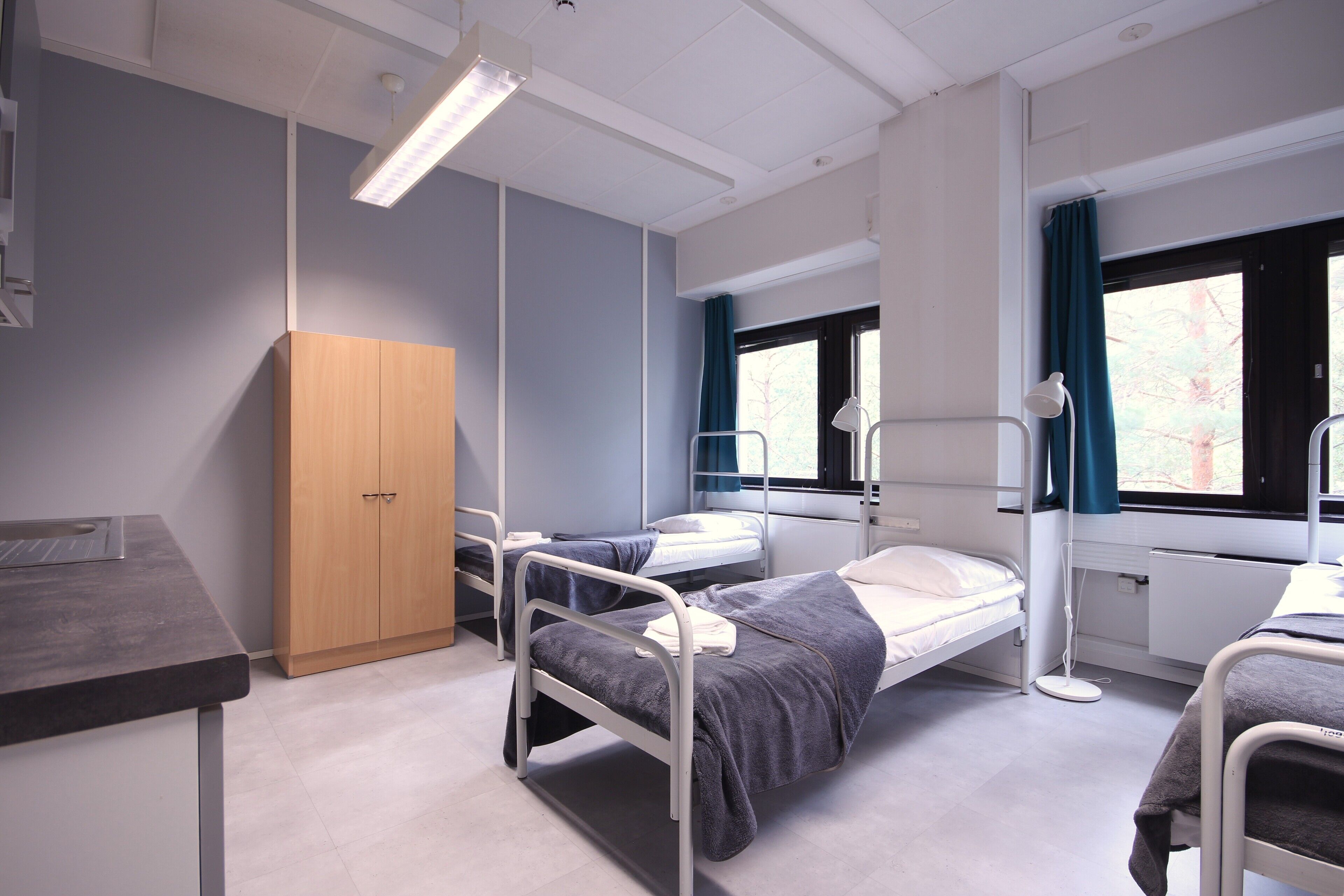 A hospital room with two beds, a desk, a cabinet, and a window with curtains.