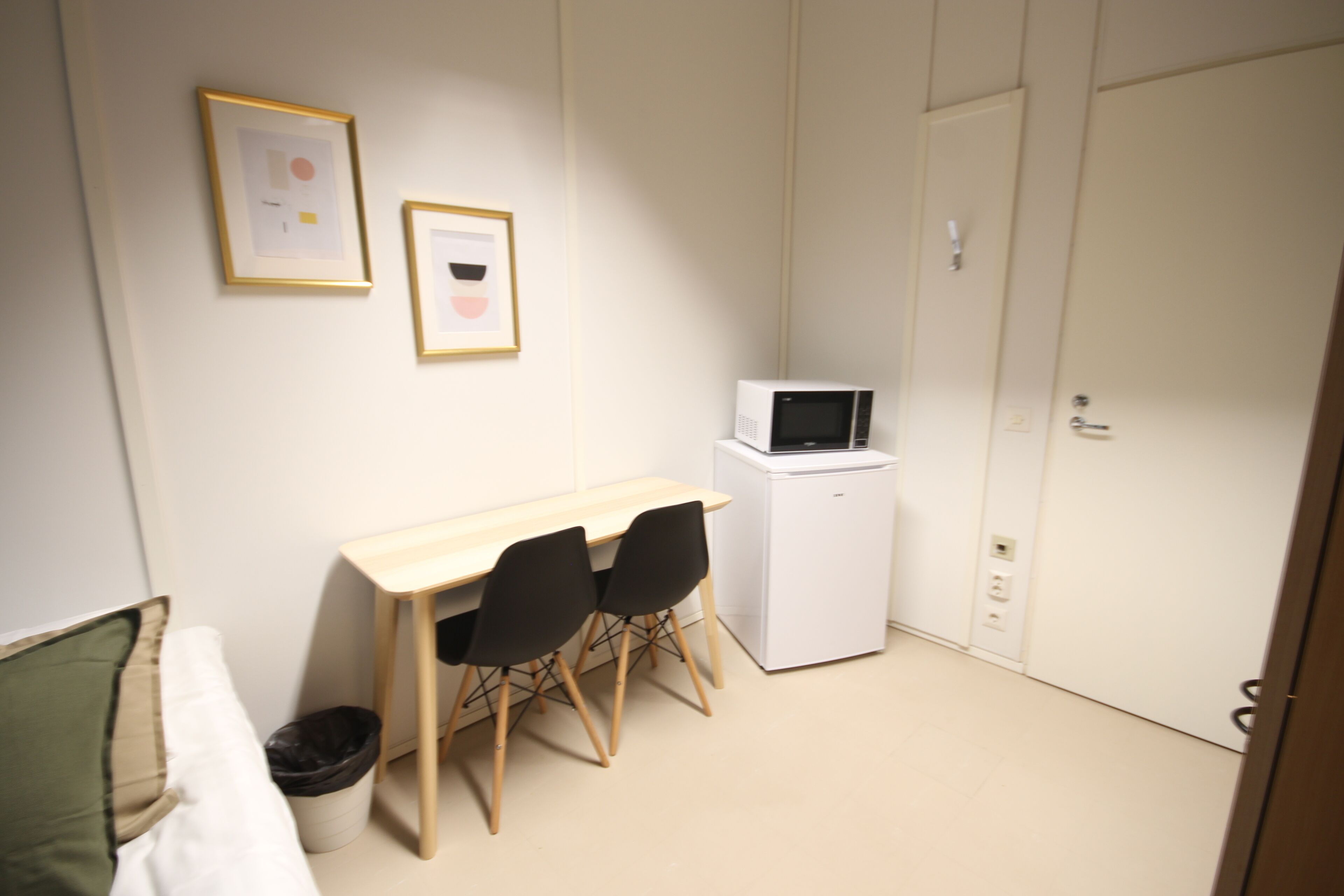 A small, compact room with a wooden desk, two black chairs, a white refrigerator, and a microwave. There is a white door and a framed picture on the wall.
