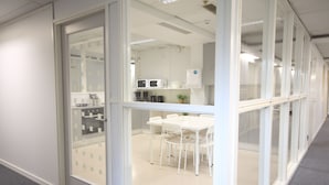Un espacio de oficina moderno con separador de vidrio, mobiliario blanco y una zona de kitchenette.
