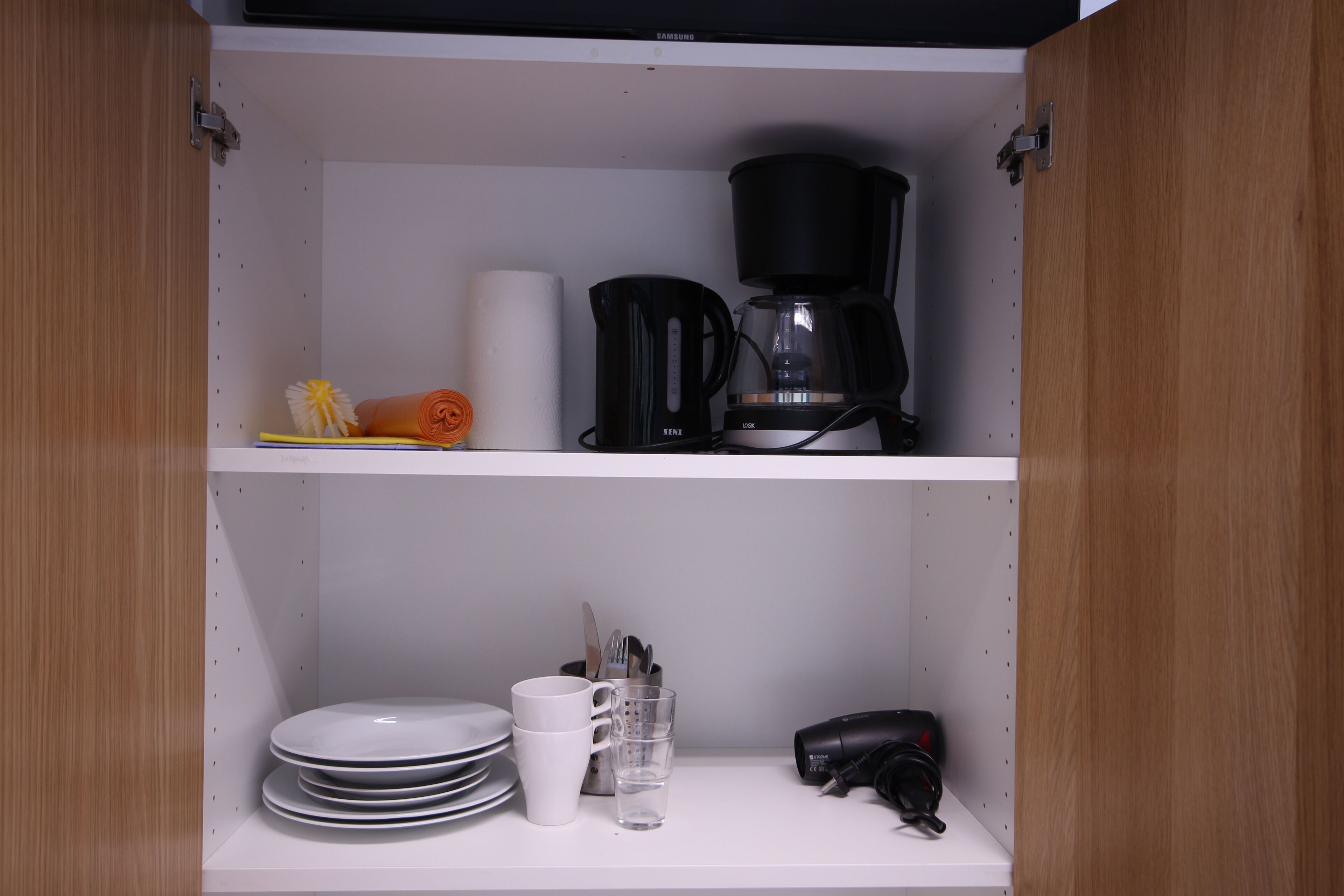 Une cuisine équipée d’une machine à café, d’assiettes, de tasses et d’un sèche-cheveux rangé sur les étagères.