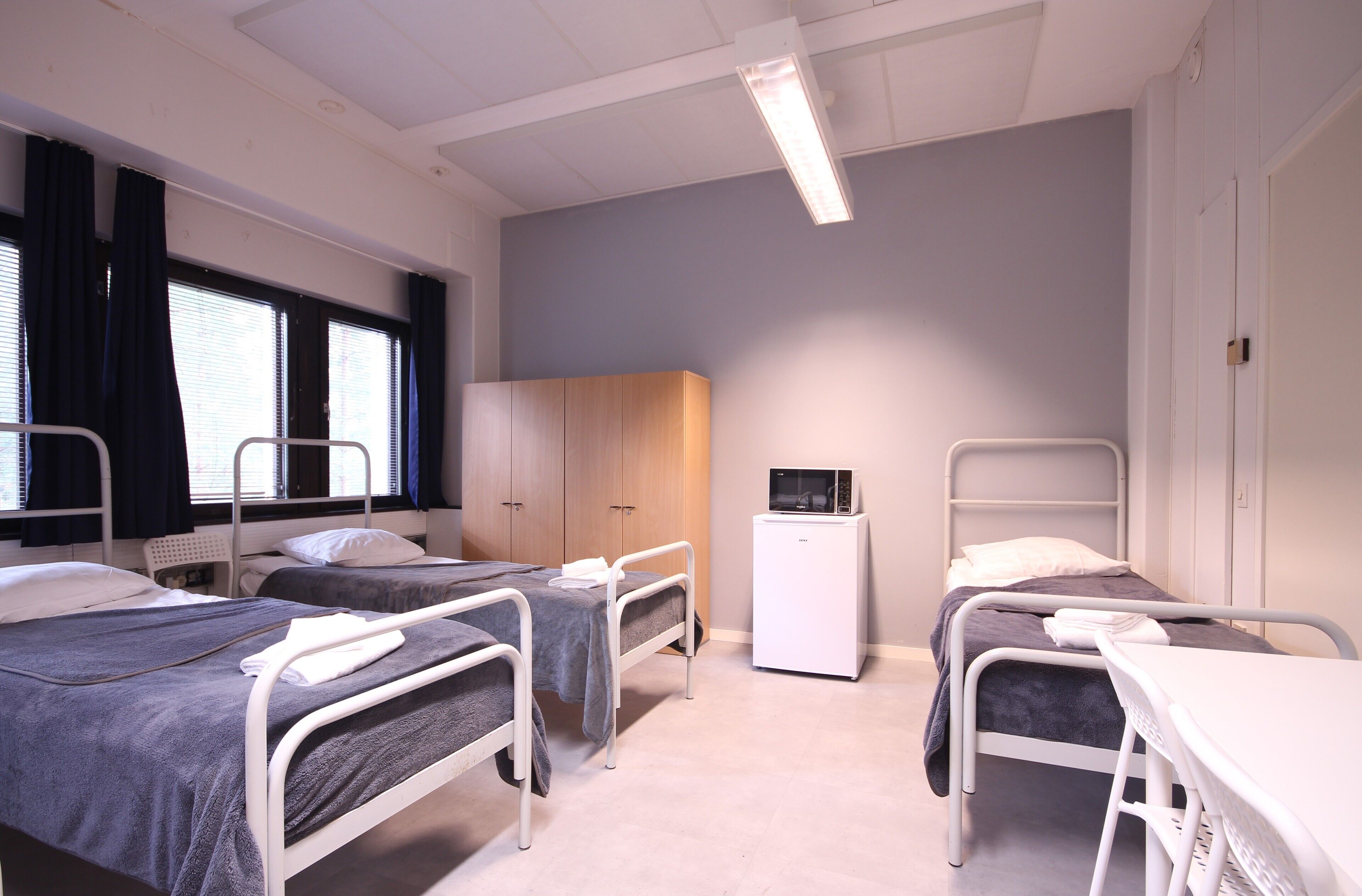 Quarto de hospital com três camas, um frigorífico branco pequeno e um guarda-roupa de madeira.