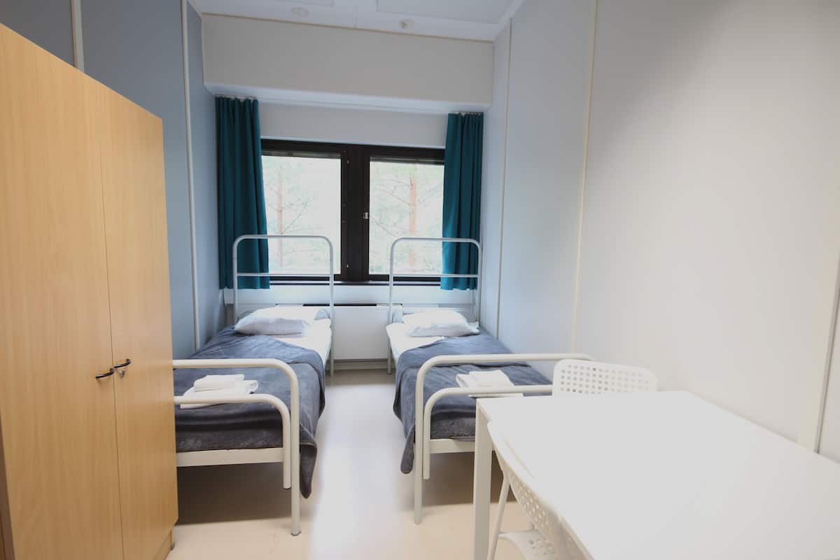 A room with two single beds, a wooden wardrobe, a window with curtains, and a white table.