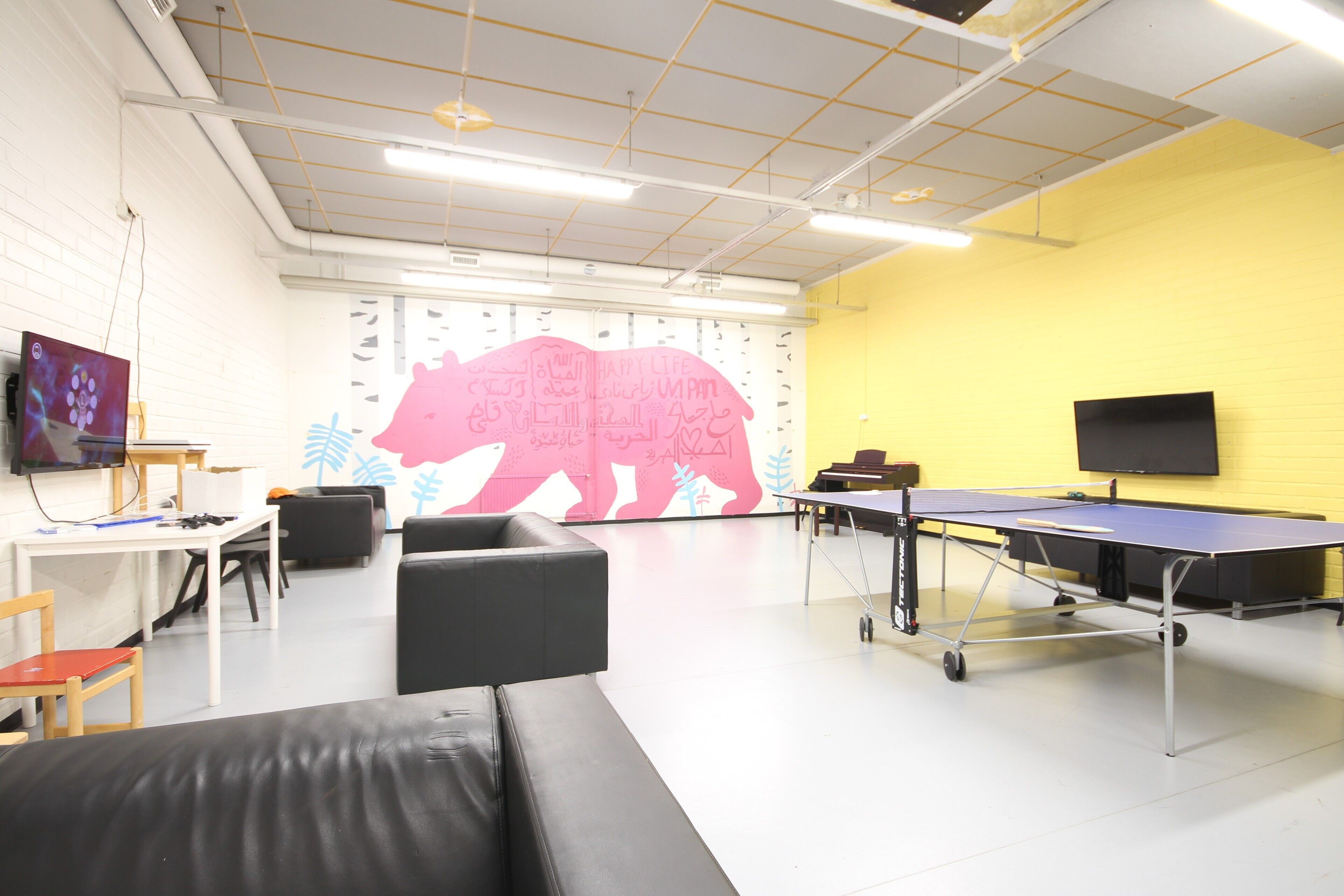 A room with a ping pong table, a sofa, a TV, and a wall with a bear mural.