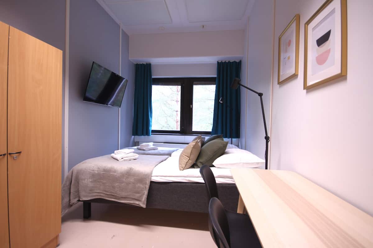 A bedroom with a bed, a desk, a chair, a TV mounted on the wall, and a window with curtains.
