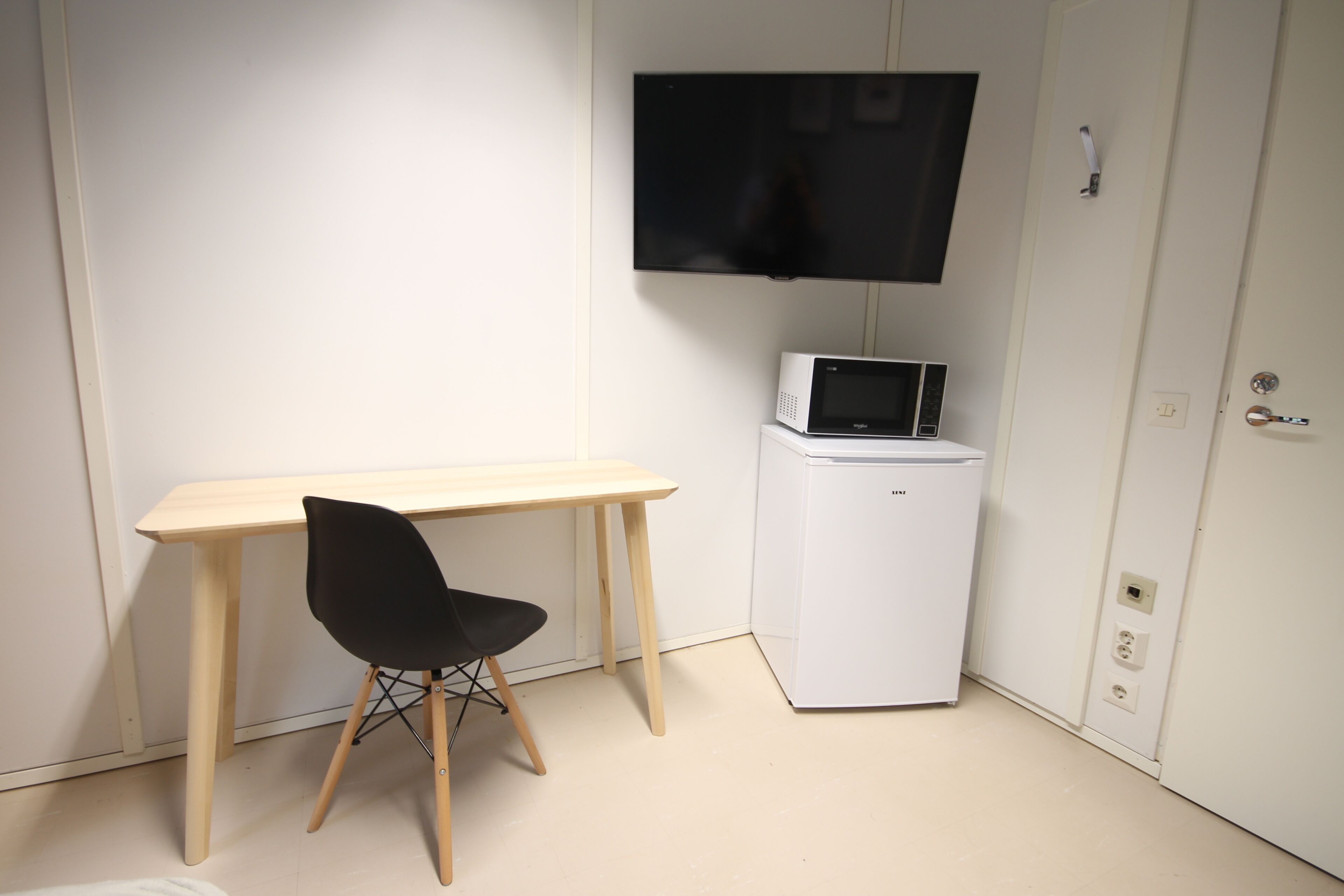 A small room with a wooden desk, a black chair, a white refrigerator, and a flat-screen TV mounted on the wall.
