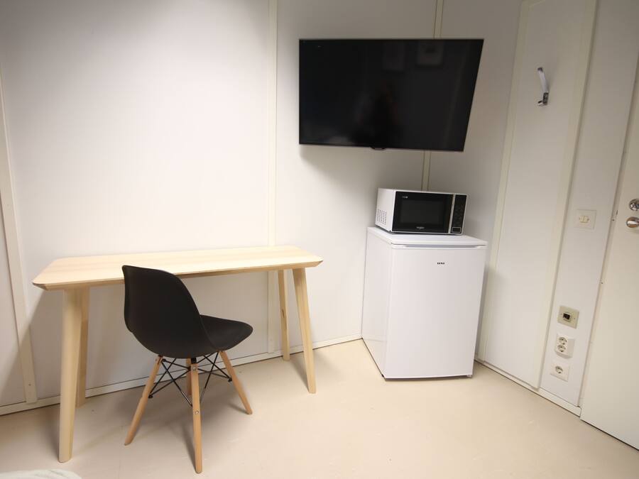 A small room with a wooden desk, a black chair, a white refrigerator, and a flat-screen TV mounted on the wall.