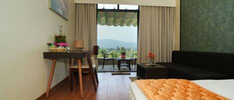 A hotel room with a bed, a desk, a chair, a small table, and a sofa. There is a window with a view of the outdoors.