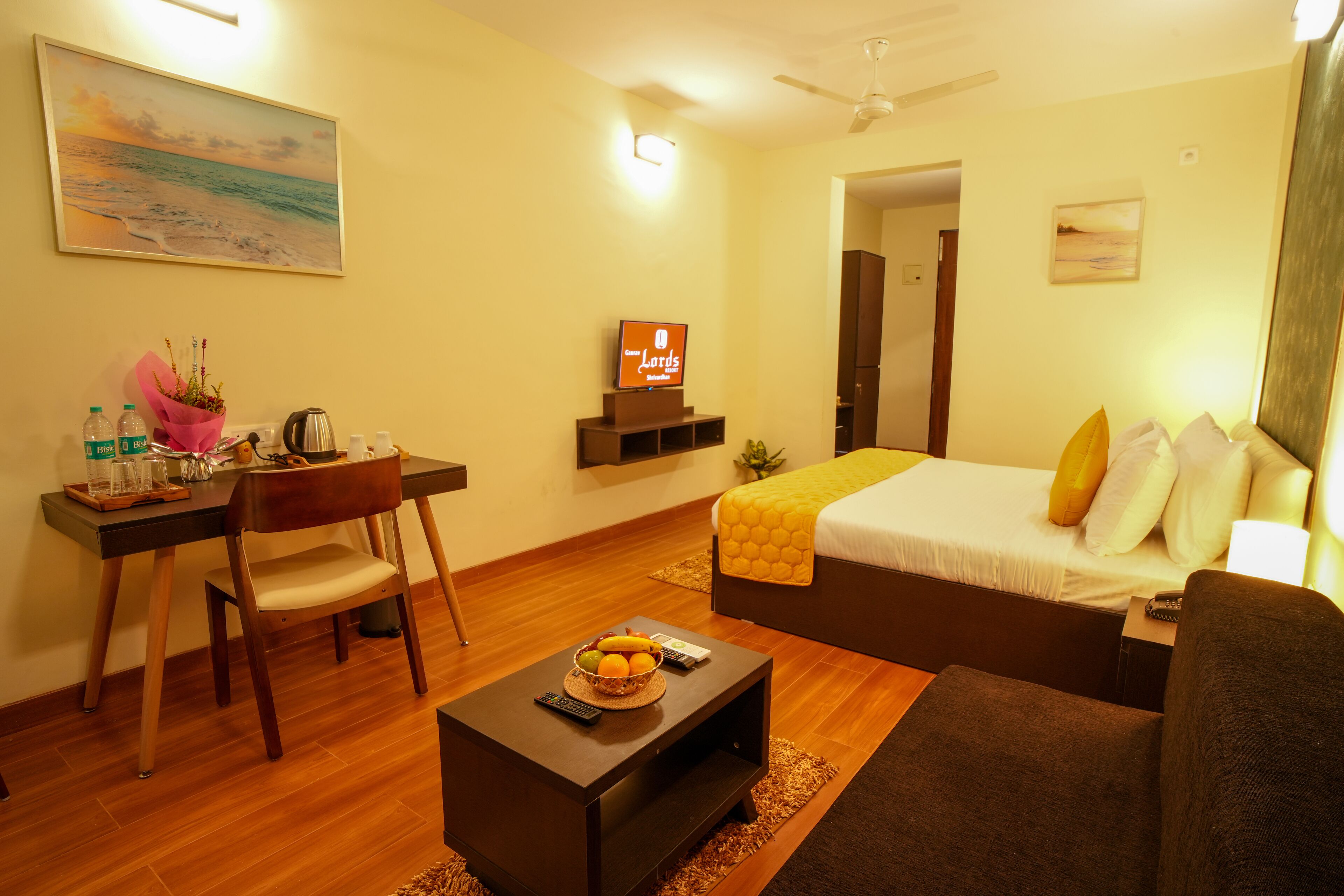 A hotel room with a bed, a desk with a chair, a coffee table, a TV, and a painting on the wall.