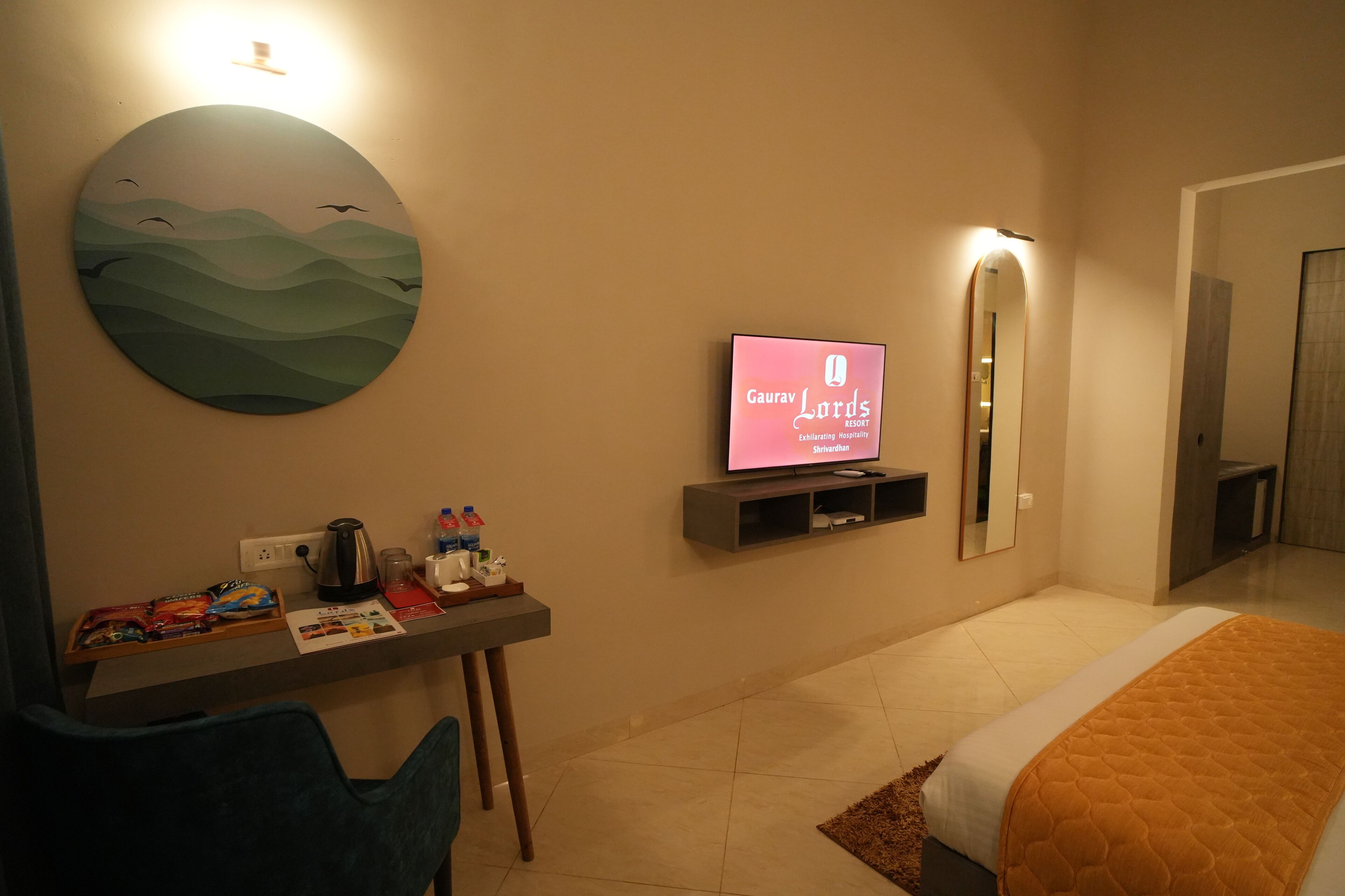 Quarto de hotel com cama, escrivaninha com cadeira, televisão, espelho e uma obra de arte na parede.