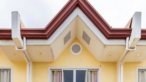 A two-story building with a yellow facade, white trim, and a red roof. There are two ground-level windows with curtains, a central door, and a small vent above the door. - WOW! 3BR House w/Karaoke ,Swimming Pool, Pet Friendly (Lapu-Lapu City)